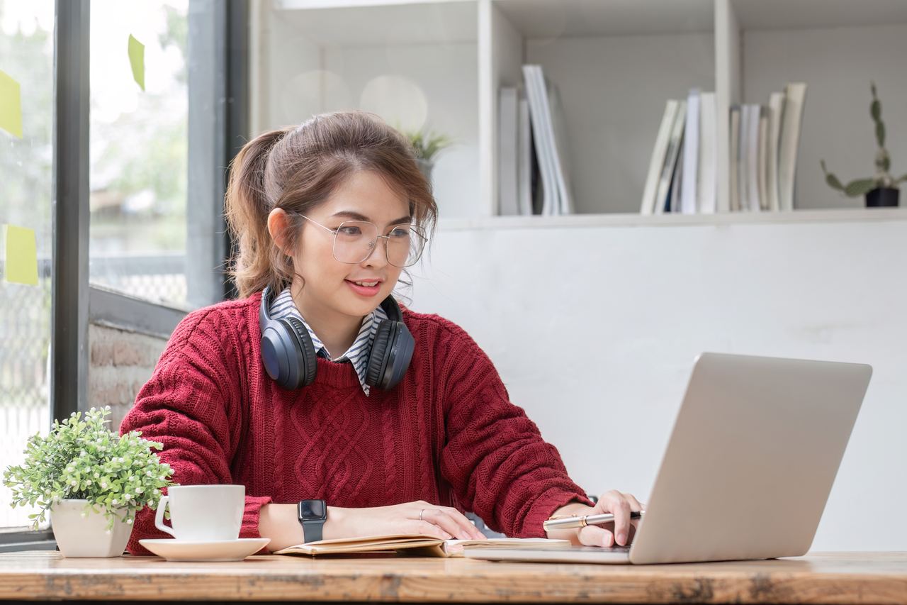 A smiling young woman with headphones around her neck studies on her laptop at a desk at home.